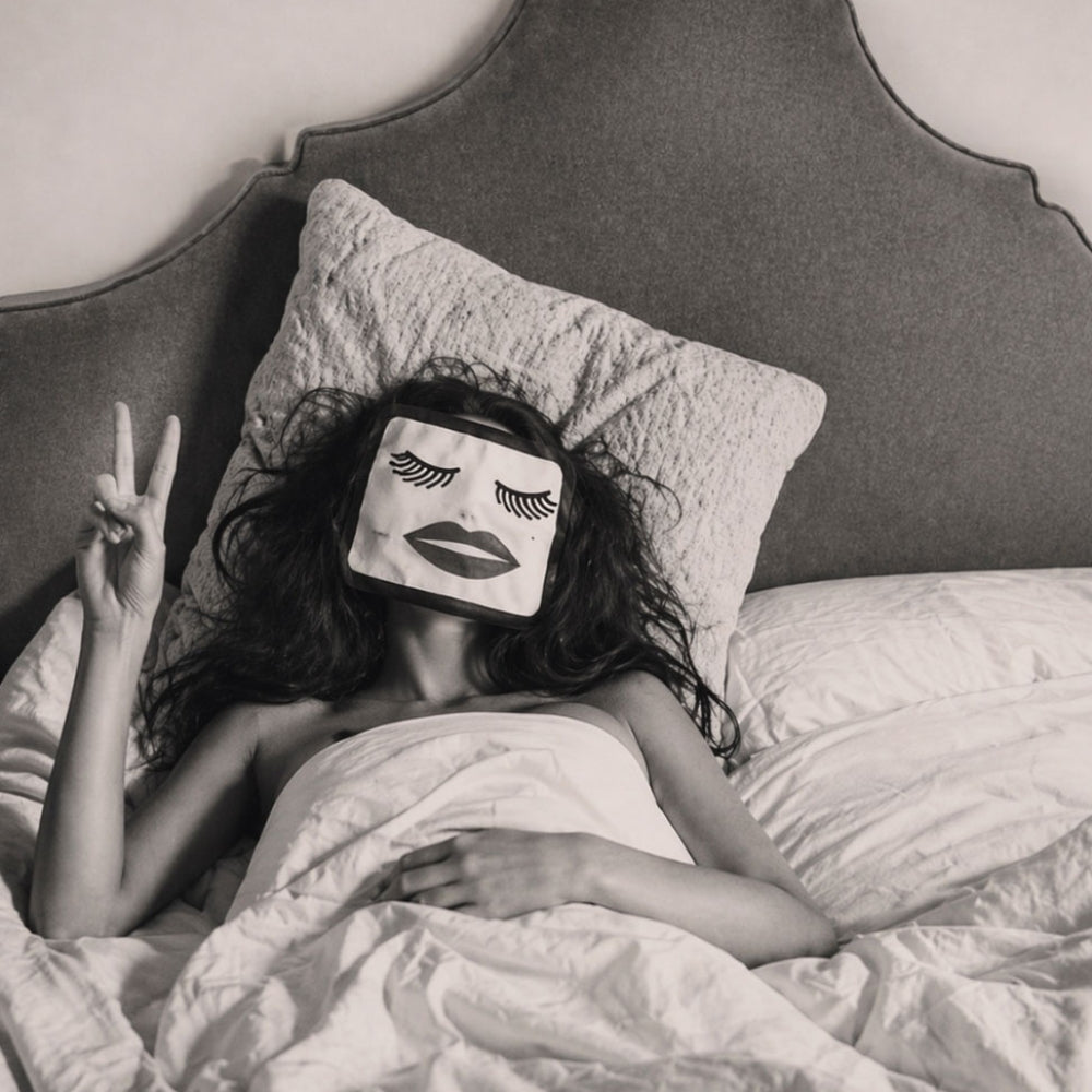 Black-and-white image of a woman in bed wearing a playful sleep mask and holding up a peace sign.