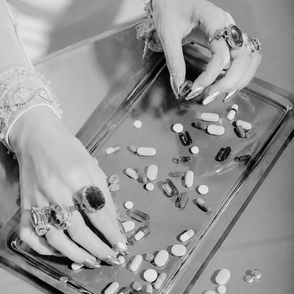 Black-and-white editorial photograph of a woman’s hands adorned with statement rings and metallic nails sorting scattered pills and supplement capsules on a silver tray, questioning the overwhelming world of perimenopause supplements and women’s wellness.